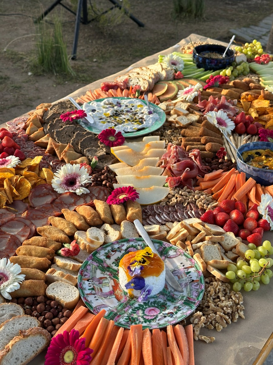 An abundant grazing table at golden hour with cured meats, cheeses, melon, breads and hand-painted plates of flower-topped cheese