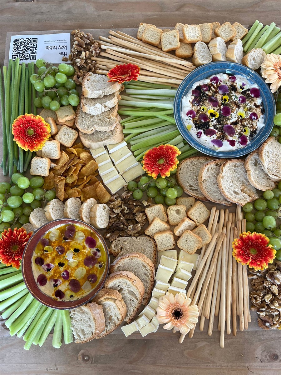Top-down board with breadsticks, dips in a flower-topped bowl, celery, grapes and gerberas beside The Lazy Table card